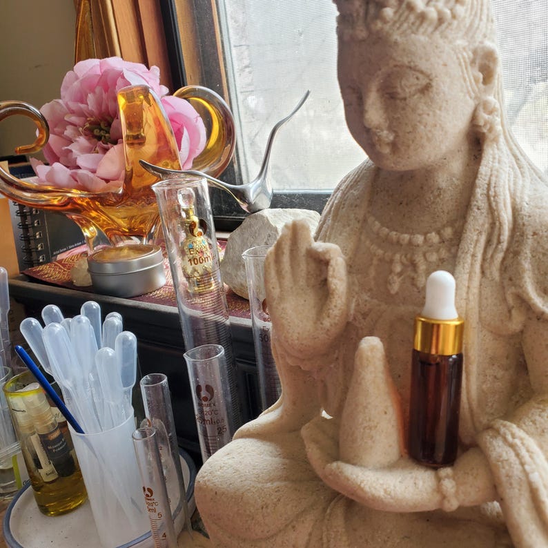 May include: A brown glass essential oil bottle with a white dropper top sits in the hand of a stone statue. The statue is of a seated figure with a serene expression. Other lab equipment and a pink flower arrangement are in the background.
