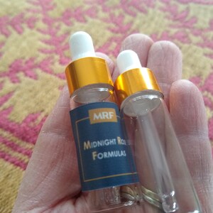 May include: Two small, clear glass bottles with white droppers and gold-colored caps are held in a hand. One bottle has a navy blue label with the text "MRF Midnight Rose Formulas". The background is a patterned fabric.