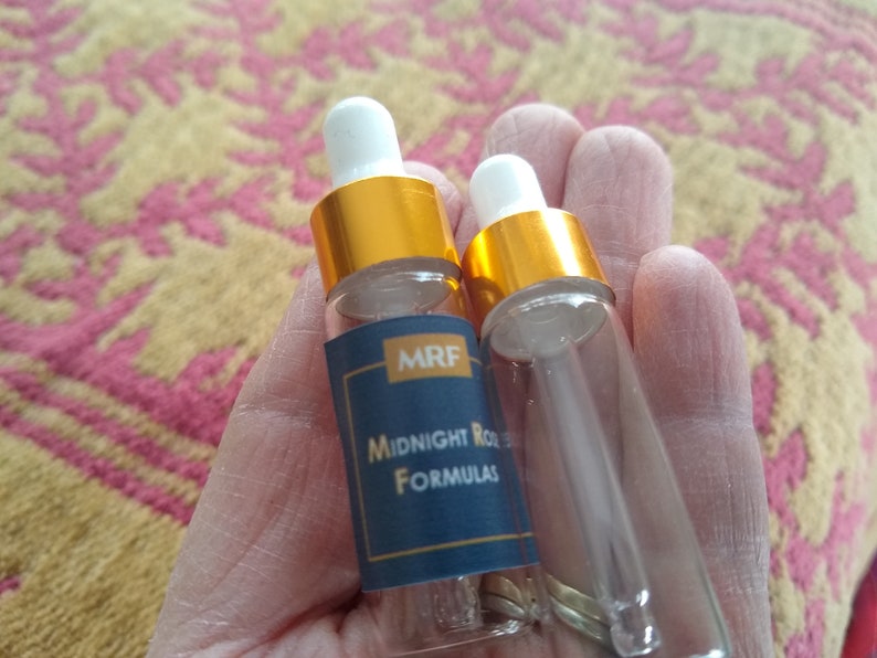 May include: Two small, clear glass bottles with white droppers and gold-colored caps are held in a hand. One bottle has a navy blue label with the text "MRF Midnight Rose Formulas". The background is a patterned fabric.