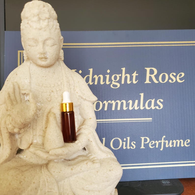 May include: A small, dark brown glass bottle with a white dropper and gold cap is held by a light-colored statue. The bottle is in front of a blue sign that reads "Midnight Rose Formulas 1 Oils Perfume."