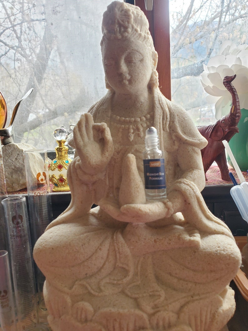 May include: A beige-colored statue of a seated figure with a serene expression, holding a small bottle. The statue has intricate details and is positioned near a window. The bottle has a blue label.