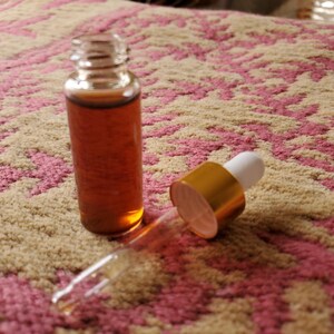 May include: A small, clear glass bottle filled with a brown liquid, next to a dropper with a gold cap and white top. The bottle and dropper are on a patterned pink and beige surface.