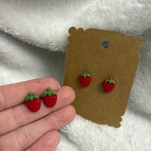 Peut inclure: Une paire de boucles d'oreilles clous en forme de fraises rouges et vertes. Les boucles d'oreilles sont présentées sur une carte marron et sur la main d'une personne.
