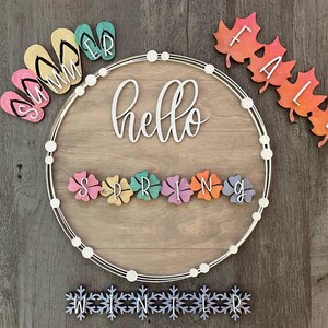 May include: A round wooden sign with the word "hello" in white lettering. The sign is decorated with colourful wooden shapes representing the seasons: flip flops for summer, flowers for spring, leaves for autumn, and snowflakes for winter.