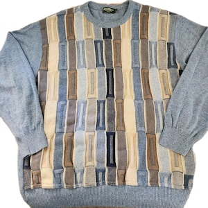May include: A light blue sweater with a geometric pattern in shades of beige, blue, and brown. The sweater has long sleeves and a crew neck. The front of the sweater features a textured, rectangular design.