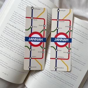 K&ouml;nnte beinhalten: Zwei rechteckige Lesezeichen mit U-Bahn-Kartendesign. Das Design zeigt einen roten Kreis mit dem Wort "JANNAH" in Blau, umgeben von farbigen Linien und arabischen W&ouml;rtern. Die Lesezeichen liegen auf einem offenen Buch.