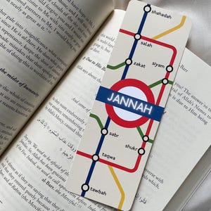 K&ouml;nnte beinhalten: Ein rechteckiger Lesezeichen mit U-Bahn-Kartendesign. Die Karte zeigt rote, blaue, gelbe und gr&uuml;ne Linien, mit Stationen, die mit islamischen Begriffen beschriftet sind. Die zentrale Station ist mit "Jannah" in wei&szlig;em Text auf einem blauen Banner beschriftet. Das Lesezeichen liegt auf einem offenen Buch.
