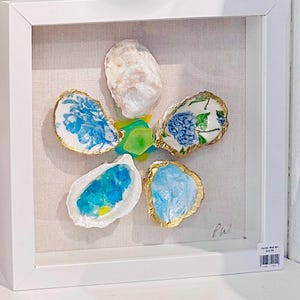 May include: A framed oyster shell art piece. The artwork features a flower design made of oyster shells with gold edges and blue and white painted interiors. The frame is white, and the background is a neutral fabric.