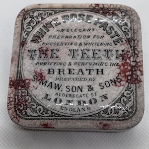 Könnte beinhalten: Rechteckige, antike Blechdose mit dekorativem Rand und dem Text "White Rose Paste". Die Dose ist als elegante Zubereitung zur Erhaltung und Aufhellung der Zähne und zur Reinigung des Atems gekennzeichnet.