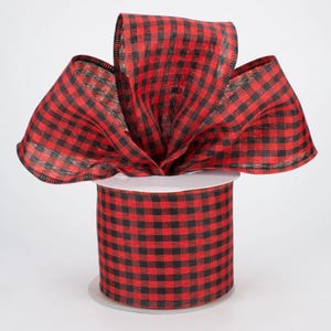 May include: A spool of wide ribbon featuring a red and black buffalo check pattern. The ribbon is wrapped around a cylindrical core, with a large, multi-looped bow extending from the top, showcasing the textured, woven fabric of the material.