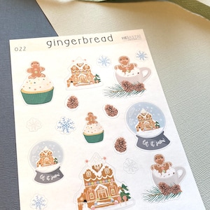Puede incluir: Una hoja de pegatinas con temática de pan de jengibre con imágenes de casas de pan de jengibre, bolas de nieve y figuras de pan de jengibre. Las pegatinas están sobre un fondo blanco y tienen una combinación de colores azul claro y marrón. El texto "Let it snow" aparece en dos de las pegatinas.