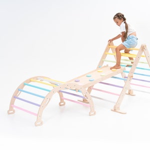 May include: A wooden climbing structure for children, featuring a triangular frame with a ladder, a ramp with colorful circles, and a curved archway with rainbow-colored rungs. A child is climbing the ladder.