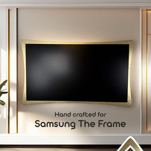 May include: A black Samsung The Frame television with a gold-colored frame is mounted on a beige wall. The text "Hand crafted for Samsung The Frame" is displayed below the TV. The room has a modern design.