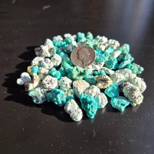 May include: A pile of turquoise stones in shades of blue and green, with a silver quarter coin for size comparison. The stones have a rough, natural texture and vary in size. The background is a dark, reflective surface.