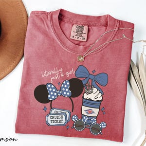 A red t-shirt with a graphic design of a cruise ticket, sunglasses, and a cup with a blue bow, along with a necklace and a hat.