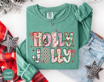 May include: Light green long-sleeved shirt with the words "HOLLY JOLLY" in a festive, patterned design. The letters are decorated with Christmas-themed patterns, bows, and stars. The shirt is folded neatly on a white surface, next to a pair of jeans.