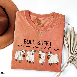 a red t-shirt with a graphic design of four cows dressed in ghost costumes, accompanied by the text "Bull Sheet" and bats. The t-shirt is placed on a white surface, with a brown hat and a fan visible in the background.