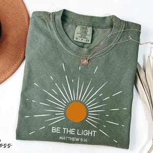 K&ouml;nnte beinhalten: Ein gr&uuml;nes T-Shirt mit einem wei&szlig;en Sonnenstrahlen-Design und dem Text "BE THE LIGHT" und "MATTHEW 5:16".