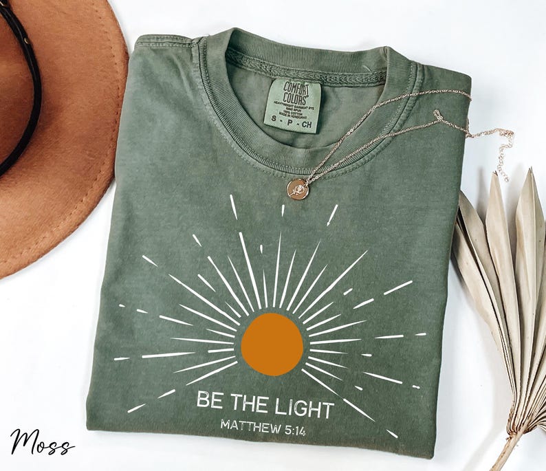 K&ouml;nnte beinhalten: Ein gr&uuml;nes T-Shirt mit einem wei&szlig;en Sonnenstrahlen-Design und dem Text "BE THE LIGHT" und "MATTHEW 5:16".