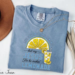 A blue t-shirt with a yellow lemon graphic and text, along with a brown hat and some dried leaves, is displayed on a white surface.