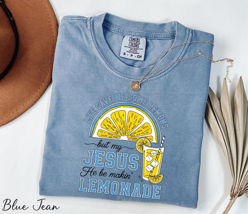 A blue t-shirt with a yellow lemon graphic and text, along with a brown hat and some dried leaves, is displayed on a white surface.