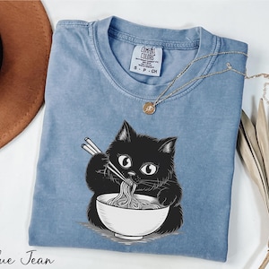 A blue denim t-shirt with a black cat illustration eating noodles, accompanied by a brown hat, a necklace, and some dried leaves.