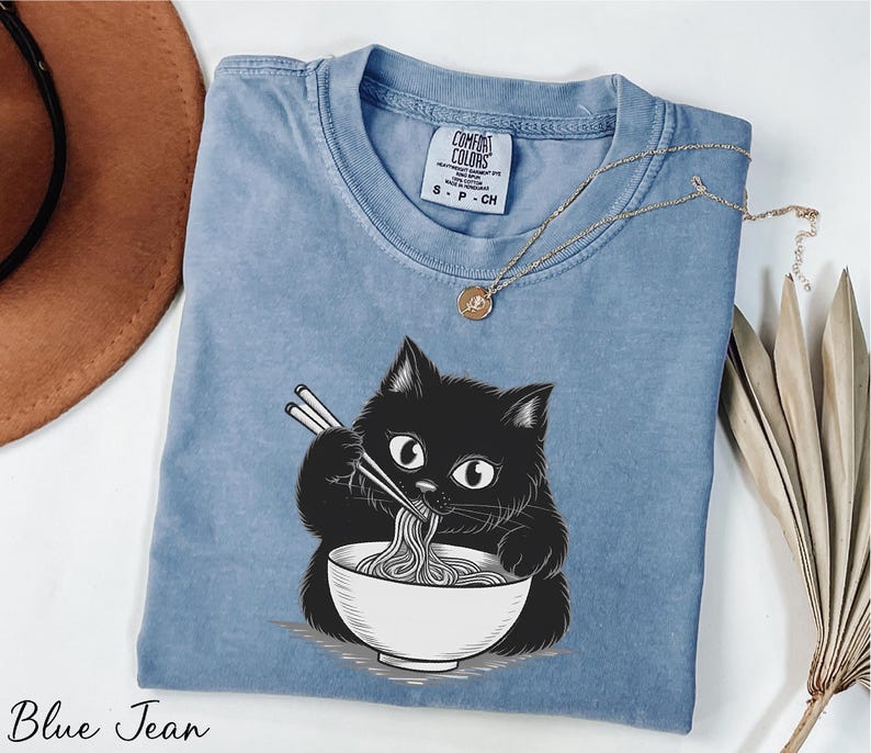 A blue denim t-shirt with a black cat illustration eating noodles, accompanied by a brown hat, a necklace, and some dried leaves.