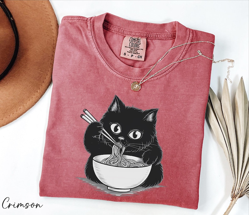 a red t-shirt with a black cat graphic, holding chopsticks and eating noodles, accompanied by a brown hat and some dried leaves.
