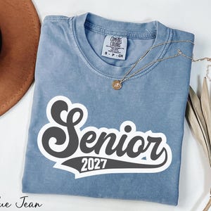 A blue t-shirt with the text "SENIOR 2027" printed on it, along with a necklace and a brown hat, all laid out on a white surface.