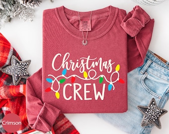 May include: Crimson long-sleeved shirt with the text "Christmas CREW" in white script, decorated with colorful string lights. A silver necklace and a pair of blue jeans are also visible. The shirt is folded, with the sleeves showing.