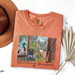 a red t-shirt with a graphic design depicting a deer, a bird, and a tree, along with the text "Hunting Season". The t-shirt is laid out on a white surface, accompanied by a hat, a necklace, and a fan.