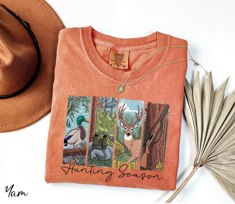 a red t-shirt with a graphic design depicting a deer, a bird, and a tree, along with the text "Hunting Season". The t-shirt is laid out on a white surface, accompanied by a hat, a necklace, and a fan.