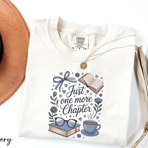 A white t-shirt with a printed design, a pair of glasses, a book, a cup of coffee, and a hat, along with a necklace and a plant, all arranged on a white background.