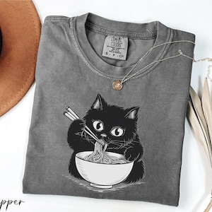 A gray t-shirt with a black cat illustration and the word "Pepper" printed on it, along with a brown hat and some dried leaves.