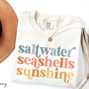 May include: Ivory t-shirt with the text "saltwater seashells sunshine" in a rainbow color scheme. The text has a vintage, distressed look.