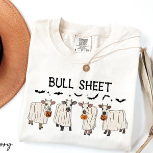 a white t-shirt with a graphic design of four cows dressed in ghost costumes, accompanied by the text "Bull Sheet" and a pumpkin. The t-shirt is placed on a white surface, with a brown hat and a fan visible in the background.