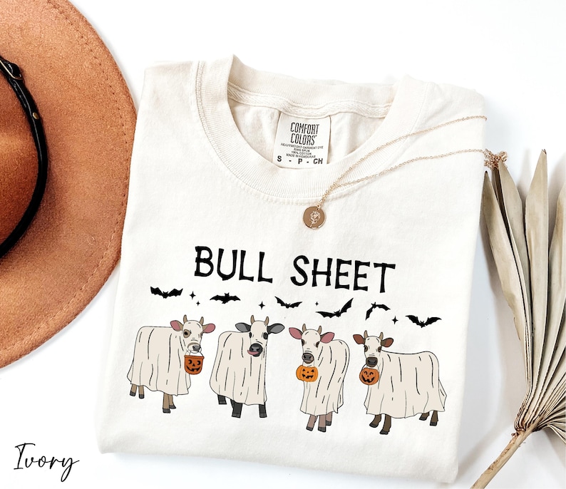a white t-shirt with a graphic design of four cows dressed in ghost costumes, accompanied by the text "Bull Sheet" and a pumpkin. The t-shirt is placed on a white surface, with a brown hat and a fan visible in the background.