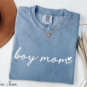 A blue t-shirt with the text "boy mom" printed on it, along with a necklace and a hat, all placed on a white surface.
