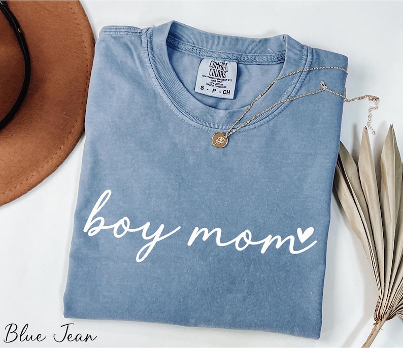 A blue t-shirt with the text "boy mom" printed on it, along with a necklace and a hat, all placed on a white surface.