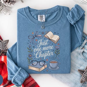 May include: A blue long-sleeve shirt with the text "Just one more chapter" and illustrations of a book, glasses, and a cup of coffee. The shirt has a crew neck and a tag that says "Comfort Colors".