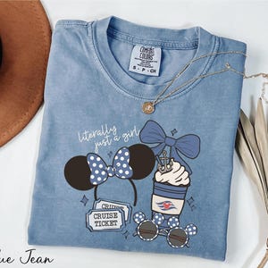 A blue denim t-shirt with a graphic design of a cruise ticket, a pair of sunglasses, and a cup with a bow, along with a necklace and a hat.