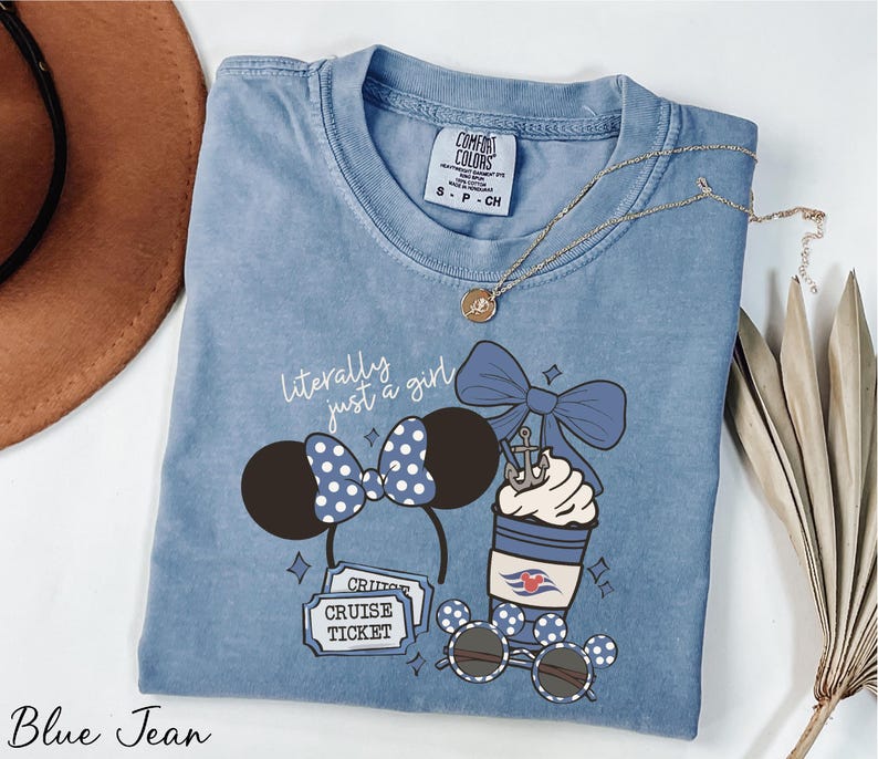 A blue denim t-shirt with a graphic design of a cruise ticket, a pair of sunglasses, and a cup with a bow, along with a necklace and a hat.