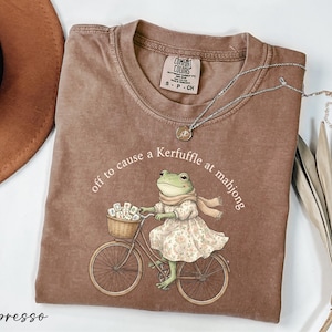 a brown t-shirt with a graphic of a frog riding a bicycle, accompanied by the text "off to cause a Kerfuffle at mahong".