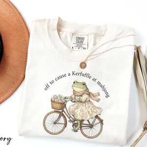 A white t-shirt with a printed illustration of a frog riding a bicycle, accompanied by the text "off to cause a Kerffuffle at mahong".