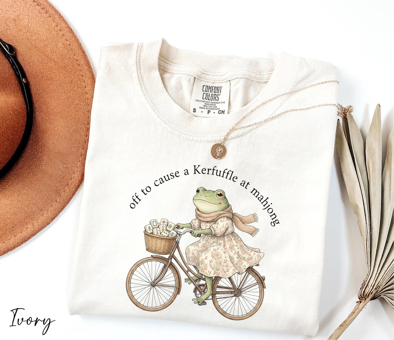 A white t-shirt with a printed illustration of a frog riding a bicycle, accompanied by the text "off to cause a Kerffuffle at mahong".