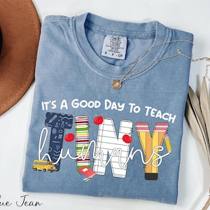 May include: A blue jean t-shirt with the text "It's a good day to teach tiny humans" in white. The text is decorated with school-themed graphics, including a school bus, pencils, apples, and books.