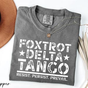May include: A gray t-shirt with a white graphic that says "FOXTROT DELTA TANGO RESIST. PERSIST. PREVAIL."