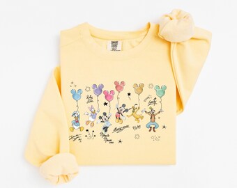 Puede incluir: Una sudadera de cuello redondo en color amarillo pálido con un estampado gráfico de personajes de Disney, incluyendo a Mickey Mouse, Minnie Mouse, Donald Duck, Daisy Duck, Pluto y Goofy sosteniendo globos de colores, con firmas y estrellas.