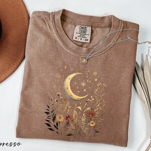 a brown t-shirt with a crescent moon and starry night sky design, along with some dried flowers and leaves. The t-shirt is placed on a white surface, and there is a brown hat and a plant in the background.