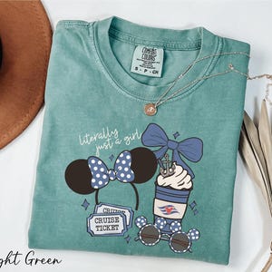 A light green t-shirt with a graphic design of a cruise ticket, a pair of sunglasses, and a cup with a bow, along with a necklace and a hat.